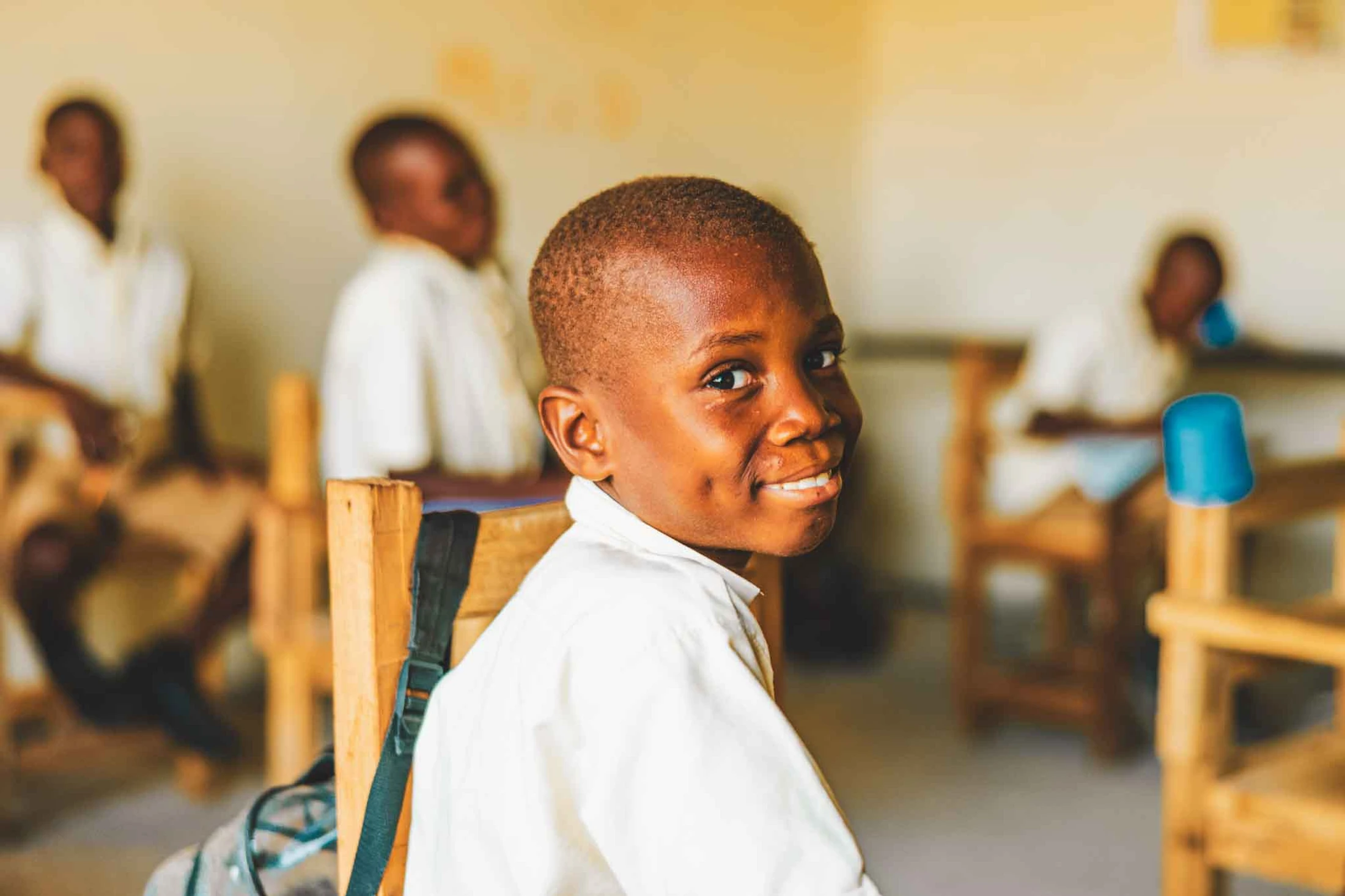 A young Black boy in a white shirt sits at a wooden desk in a classroom, looking back at the camera with a big, happy smile showing his dimples.