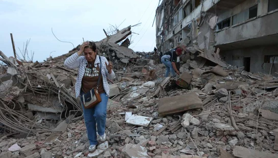 A distressed woman with her hand on her head walks through the rubble of a collapsed building. In the background, another person searches through the debris.