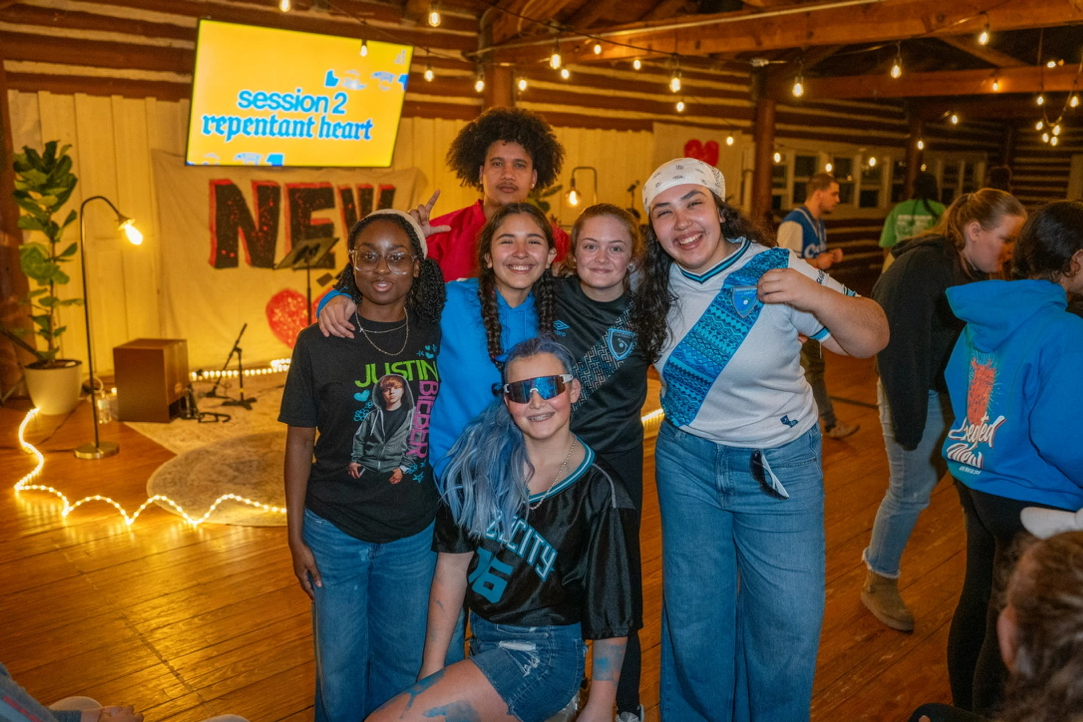 A group of six diverse young people smile for a photo in a rustic room with wood floors and walls, lit by warm string lights.