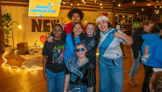 A group of six diverse young people smile for a photo in a rustic room with wood floors and walls, lit by warm string lights.
