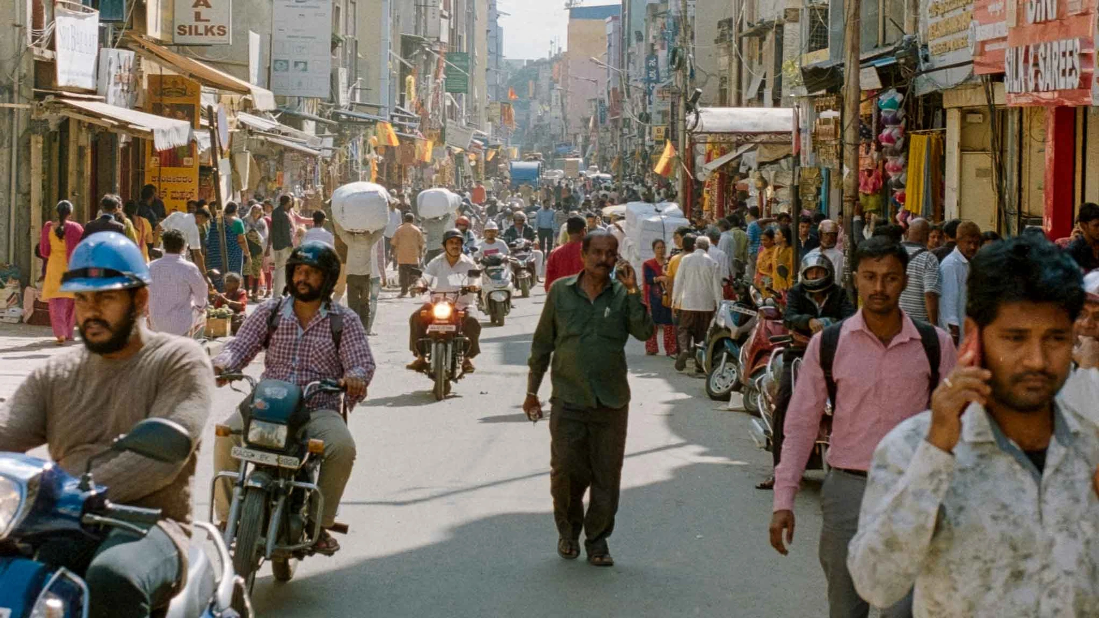 A sunlit, crowded street in an Indian city is filled with people walking and riding motorcycles between tall buildings with shops and signs.