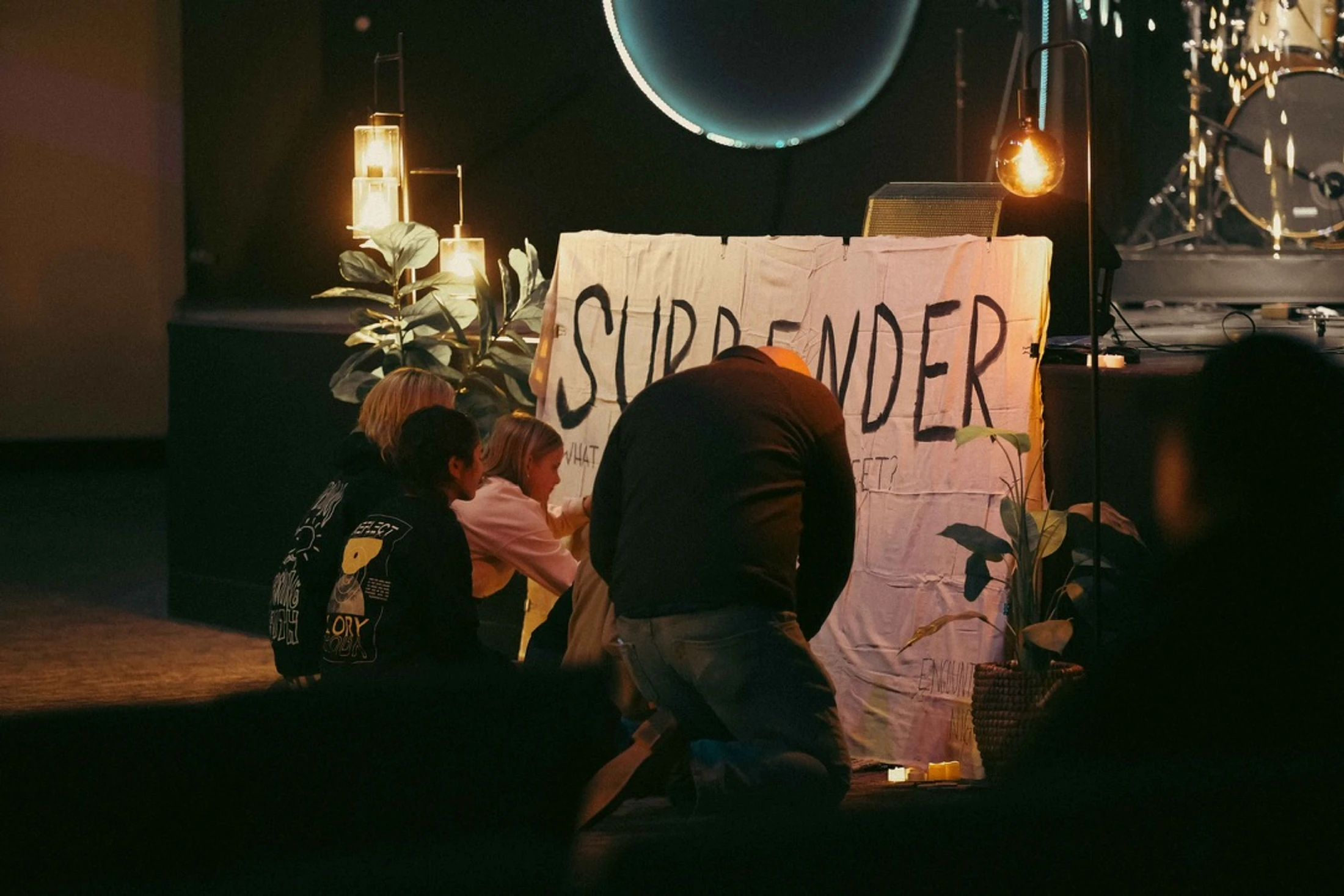 A group of people, including children, kneel on a dimly lit stage before a large, handmade sign that says 'SURRENDER' in black letters.