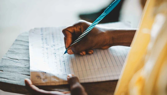 A close-up view of a person's dark-skinned hand holding a blue and white pen, writing on a piece of lined paper that rests on a wooden desk.