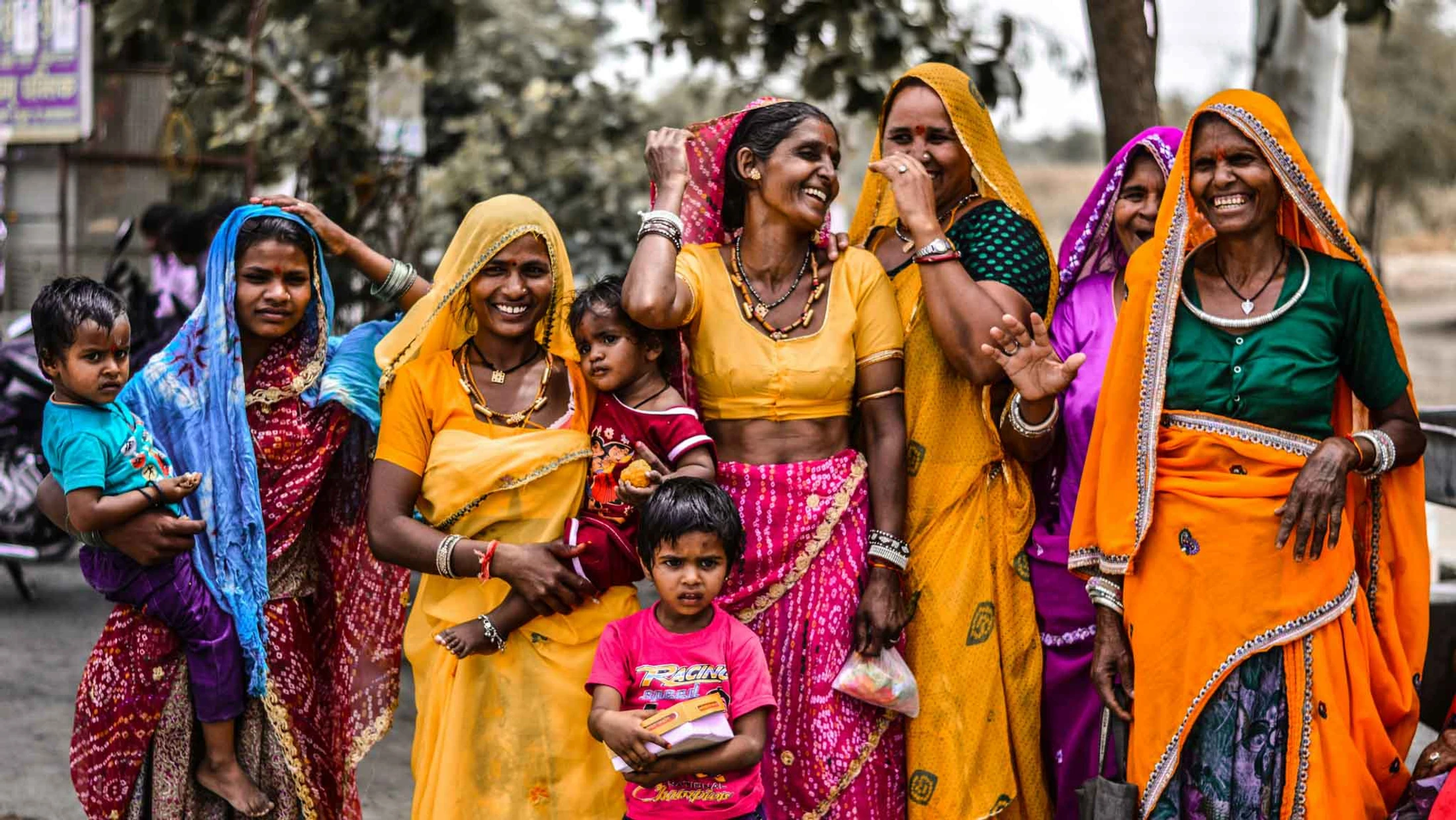A group of smiling Indian women and several children dressed in colorful traditional sarees stand together outdoors, laughing and looking at the camera.