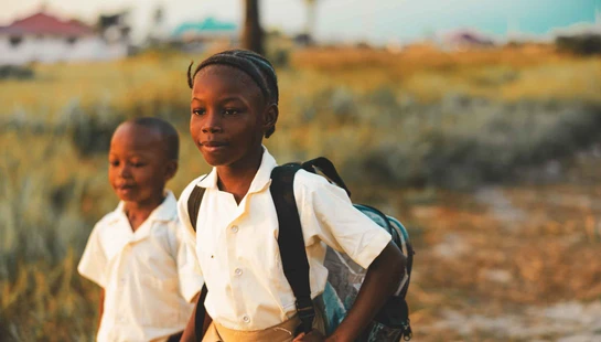 Two young Black children in school uniforms walk through a grassy field. The girl in front carries a backpack and looks ahead, illuminated by golden light.