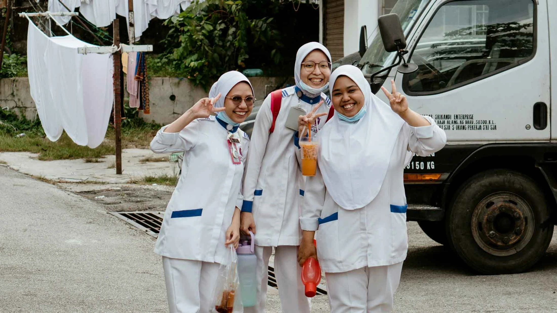 Three young women in white nursing uniforms and hijabs smile and make peace signs while posing for a photo outdoors next to a truck.
