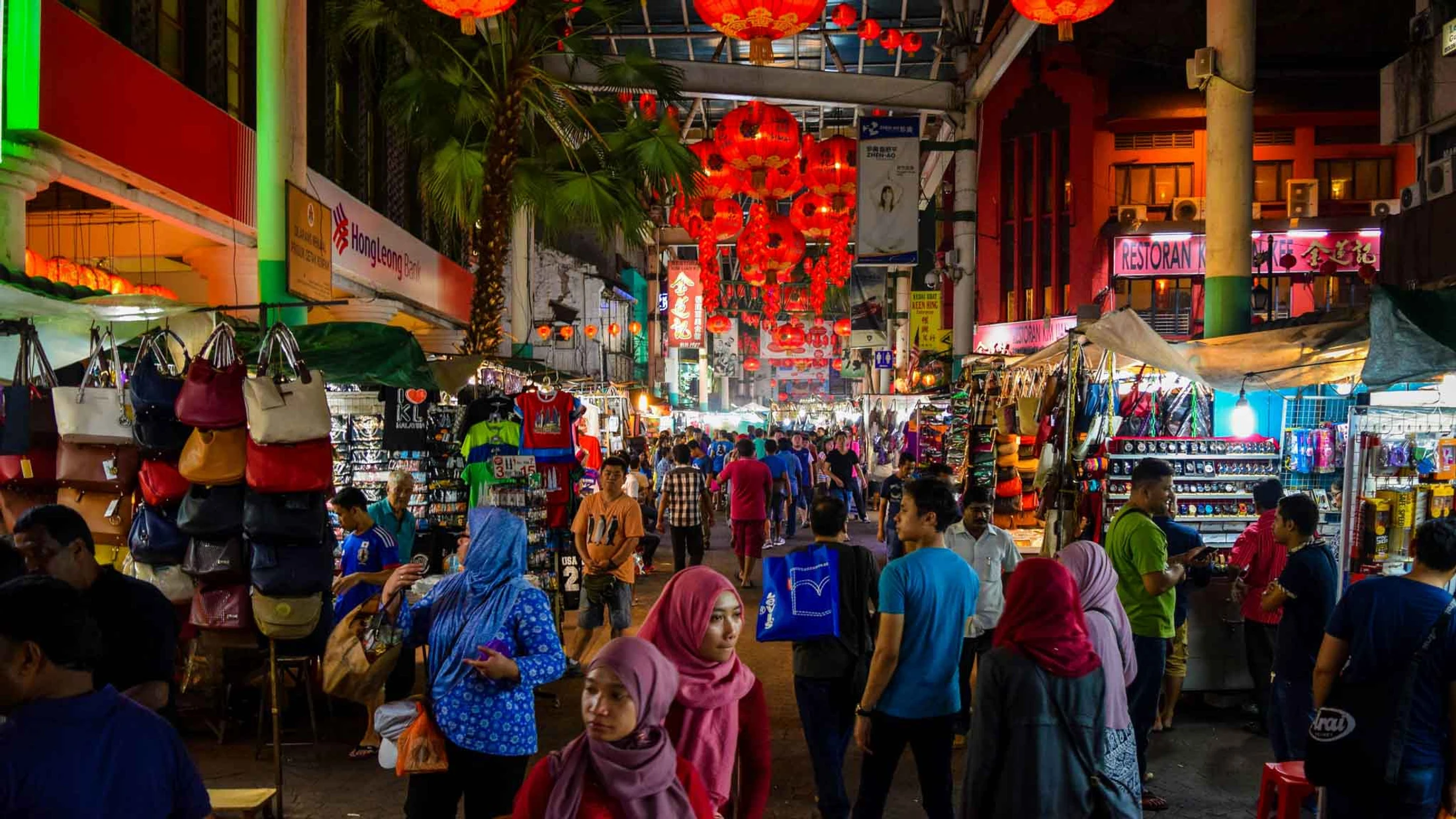 A busy street market at night in Kuala Lumpur's Chinatown, packed with people under a canopy of glowing red Chinese lanterns and market stalls.