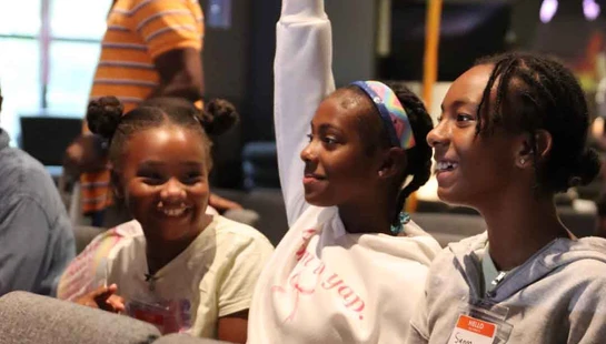 Three young Black girls sit in an audience. One girl raises her hand to answer a question, while the other two smile and look on attentively.