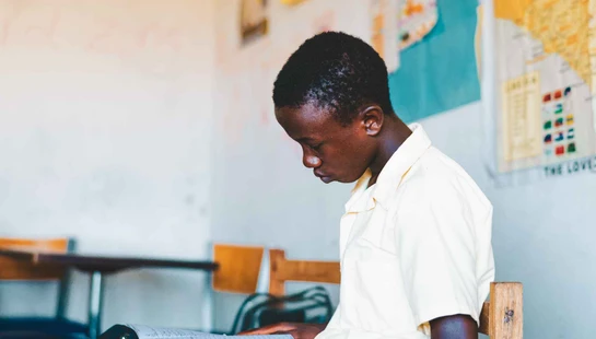 A side profile of a young Black boy in a light-colored shirt, sitting in a classroom and concentrating on a notebook he is holding.