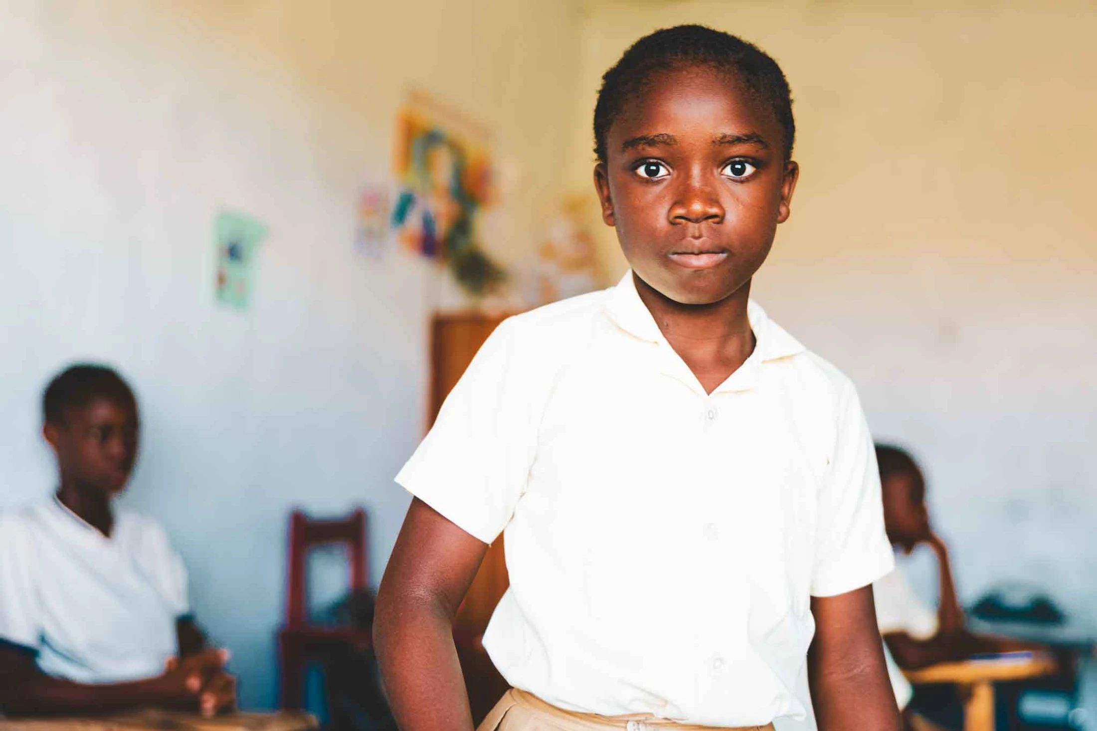 A young Black child wearing a white school uniform stands in a classroom and looks seriously into the camera. Other students are blurred in the background.