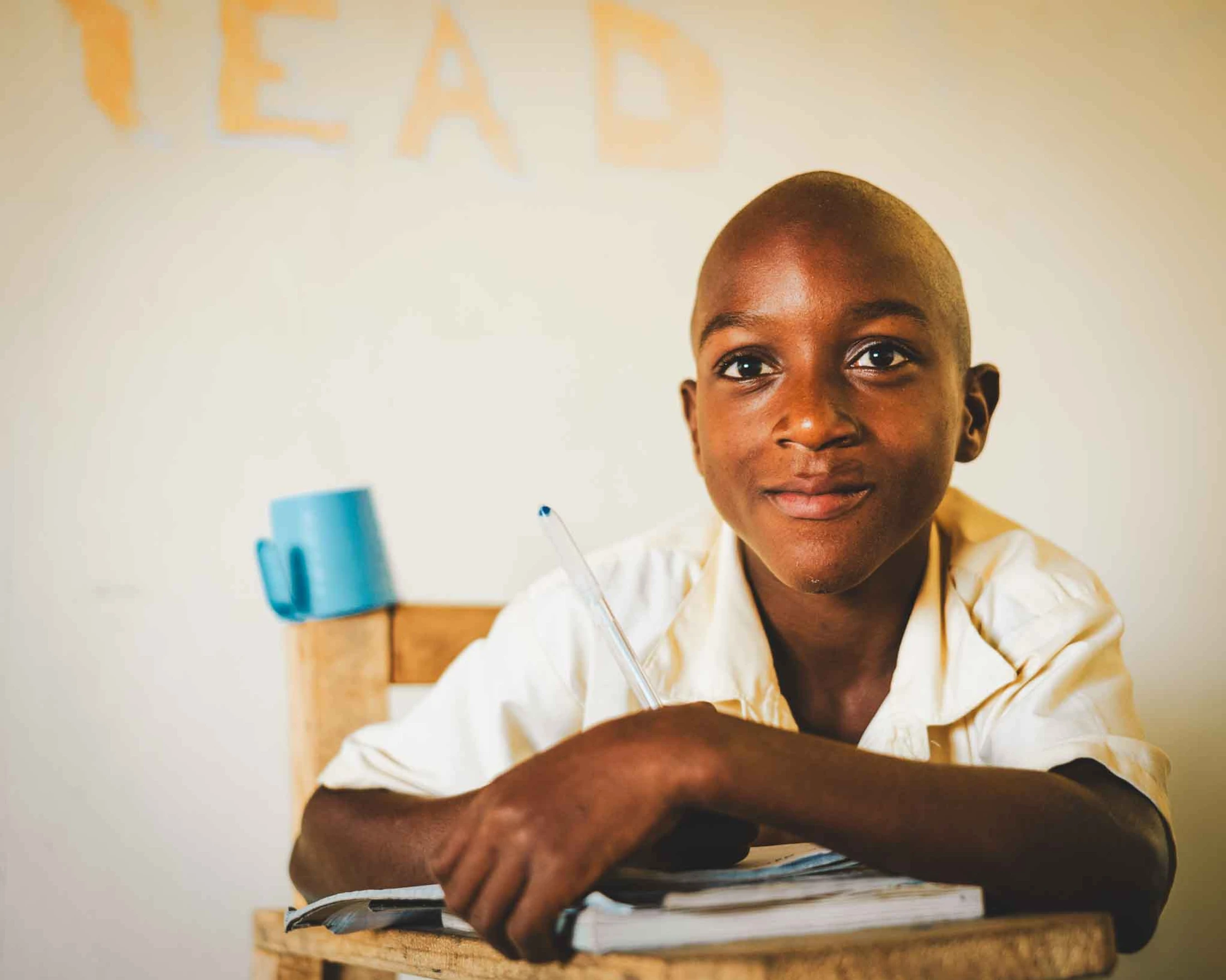 A young Black boy with a shaved head smiles warmly at the camera while sitting at a wooden desk. He holds a pen over a book, ready to learn in a classroom.