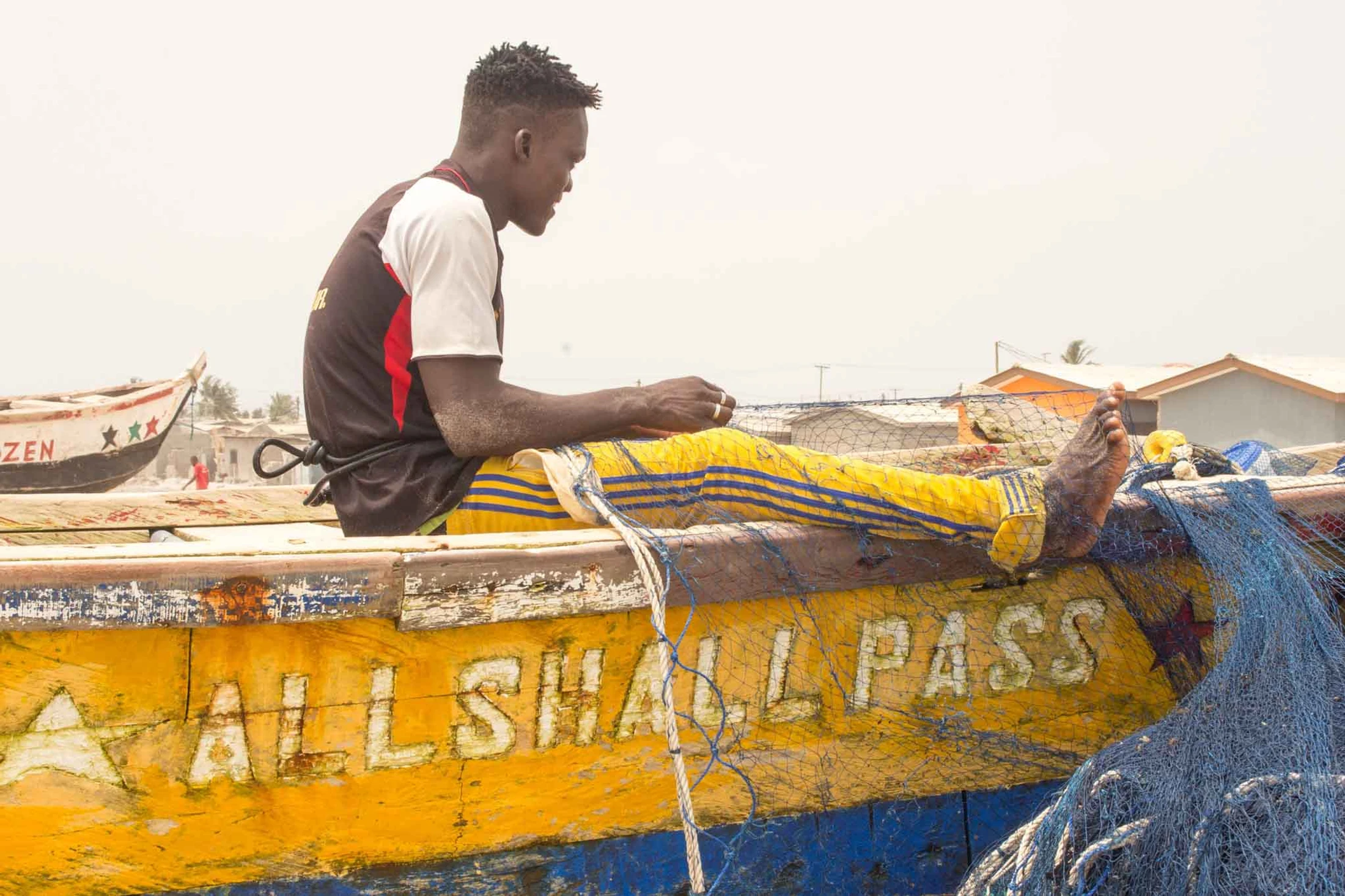 A young man sits in a weathered yellow fishing boat mending a blue net. On the side of the boat, the words 'ALL SHALL PASS' are painted in white.