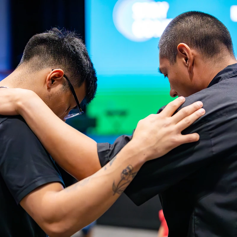 Two men with short dark hair embrace while praying. One man rests his hand on the other's shoulder in a gesture of support in front of a blue screen.