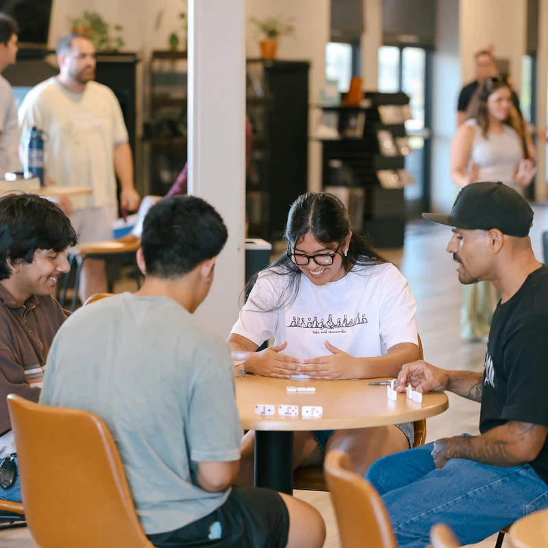 Four young adults sit around a circular table, smiling and laughing as they play a game of dominoes in a bustling common area.