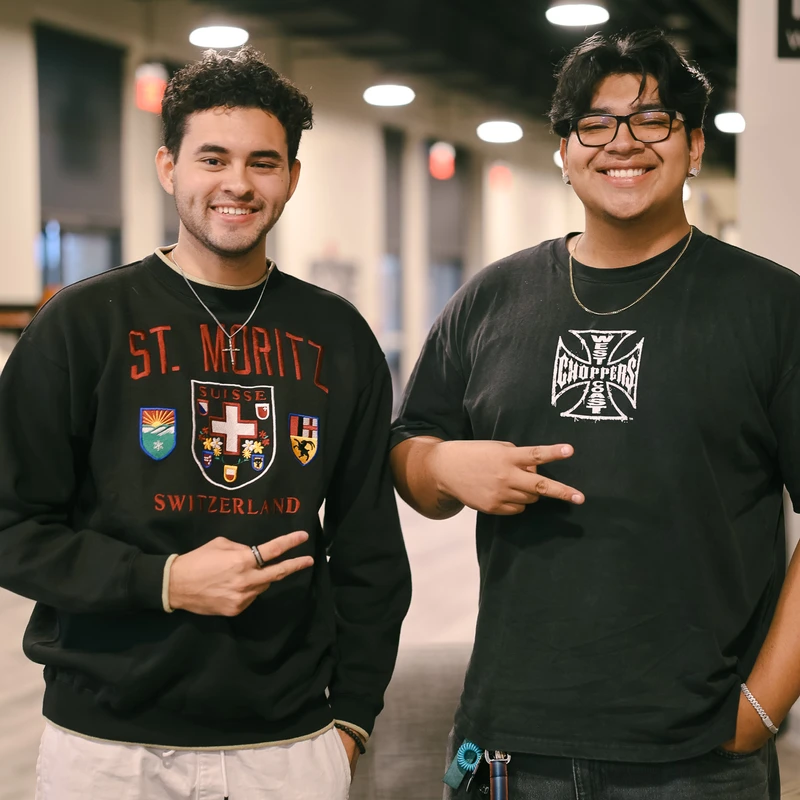 Two young men stand side-by-side in a hallway, smiling at the camera and making peace signs with their hands. They are dressed in casual clothing.