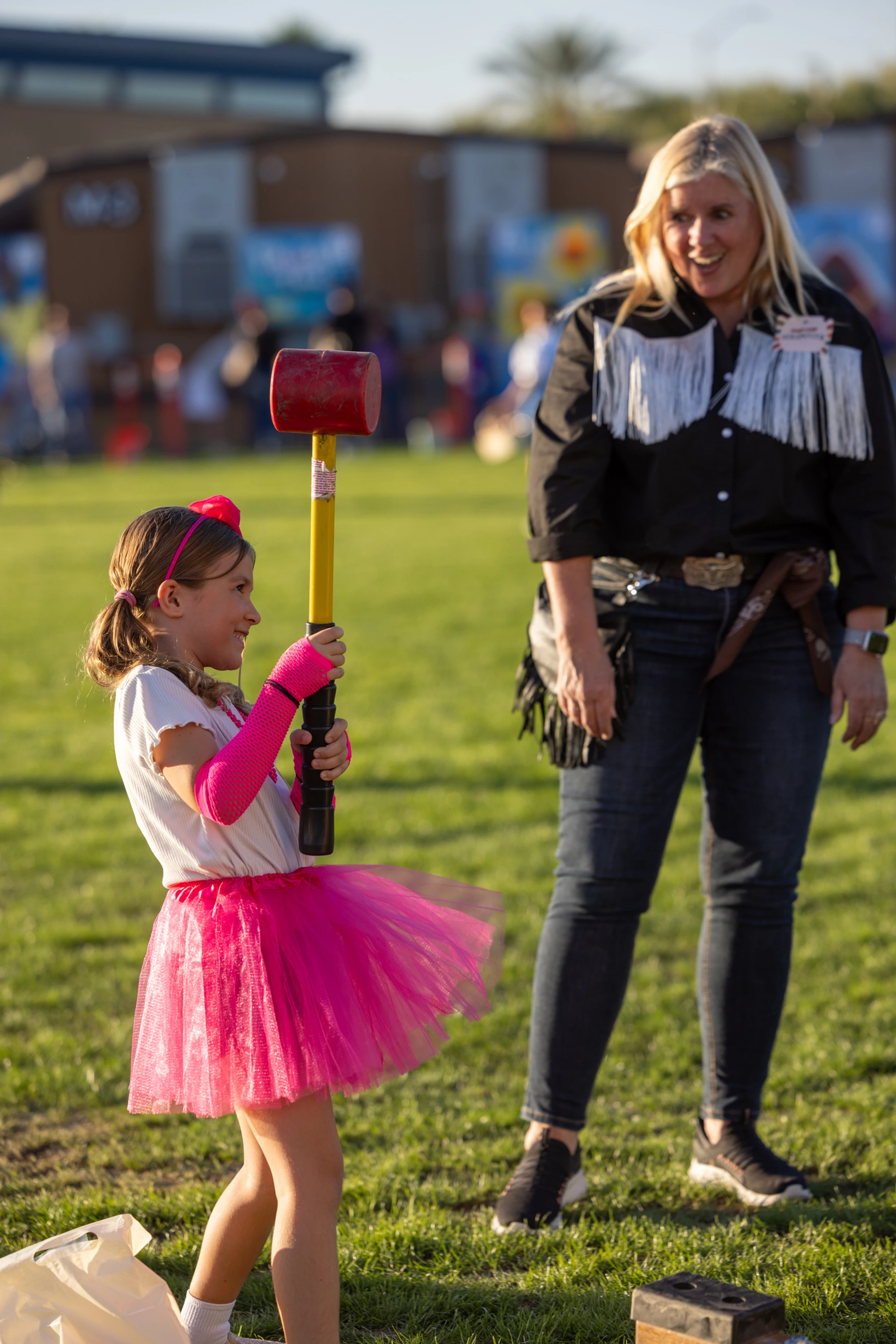 A young girl in a pink tutu smiles as she swings a large mallet at a carnival game, while a woman in a western shirt watches with a happy expression.