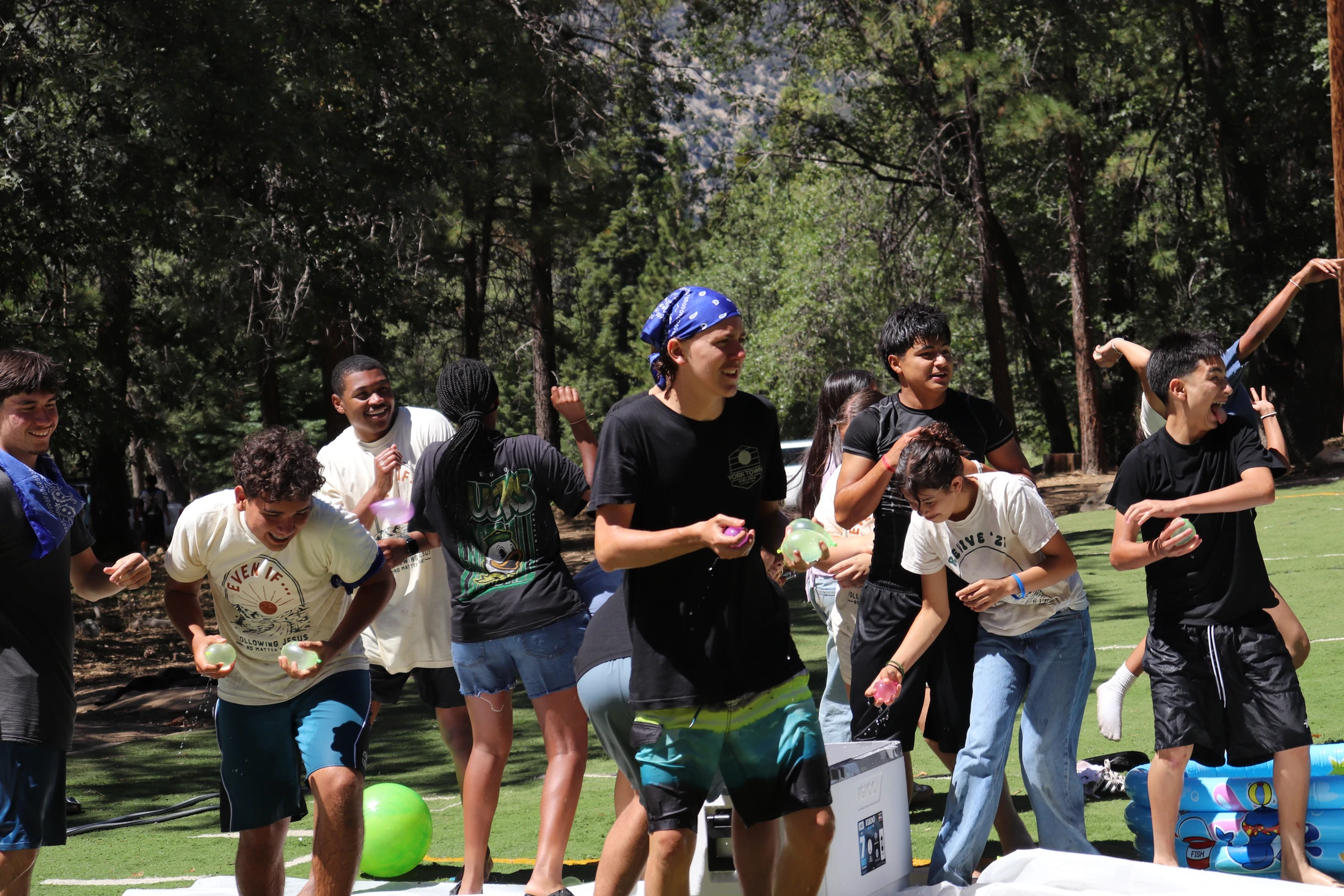 A diverse group of teenagers laughing and playing with water balloons on a green turf field, with a forest in the background on a sunny day.