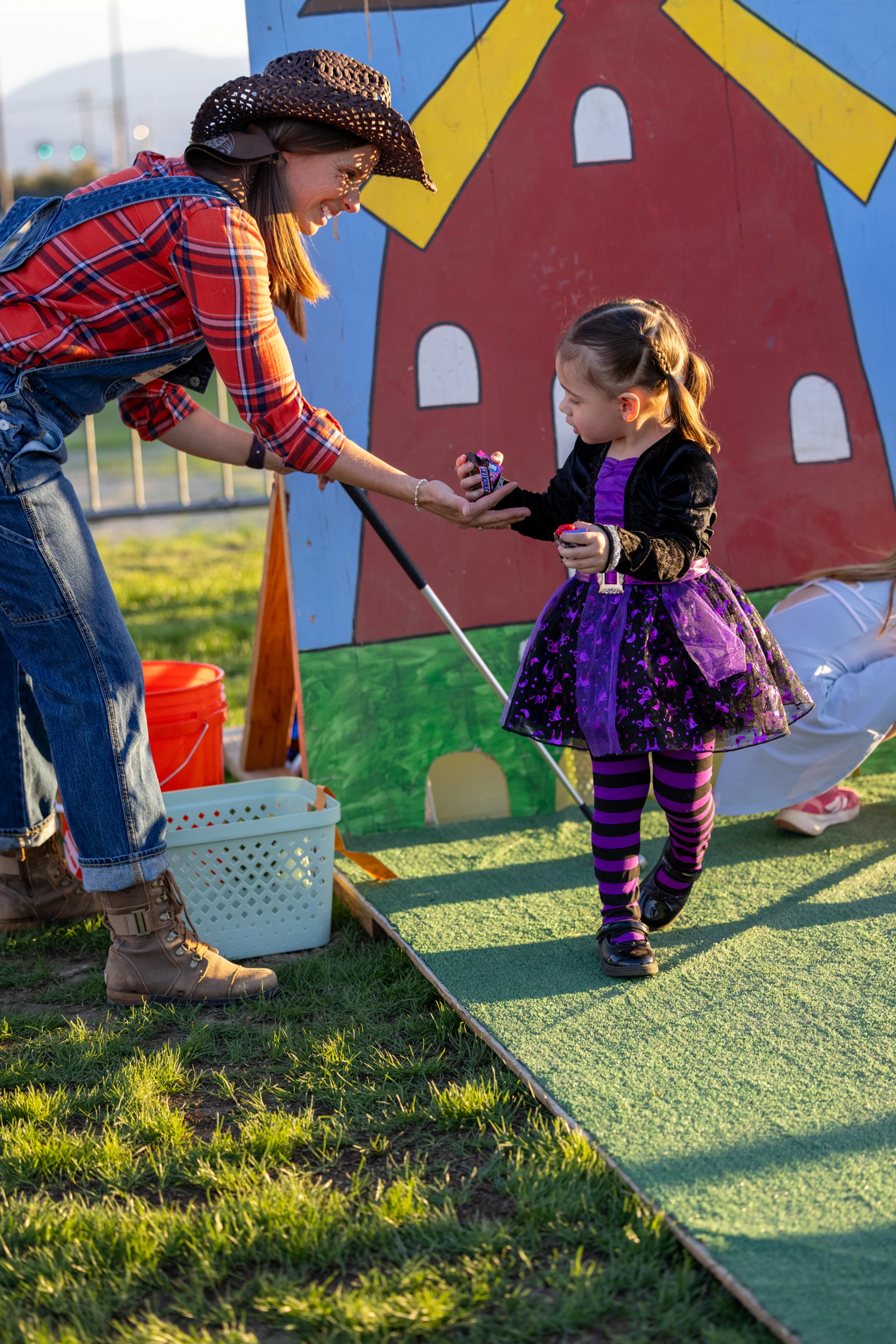 A woman dressed as a farmer gives candy to a little girl in a purple and black witch costume at an outdoor Halloween event with a painted barn backdrop.
