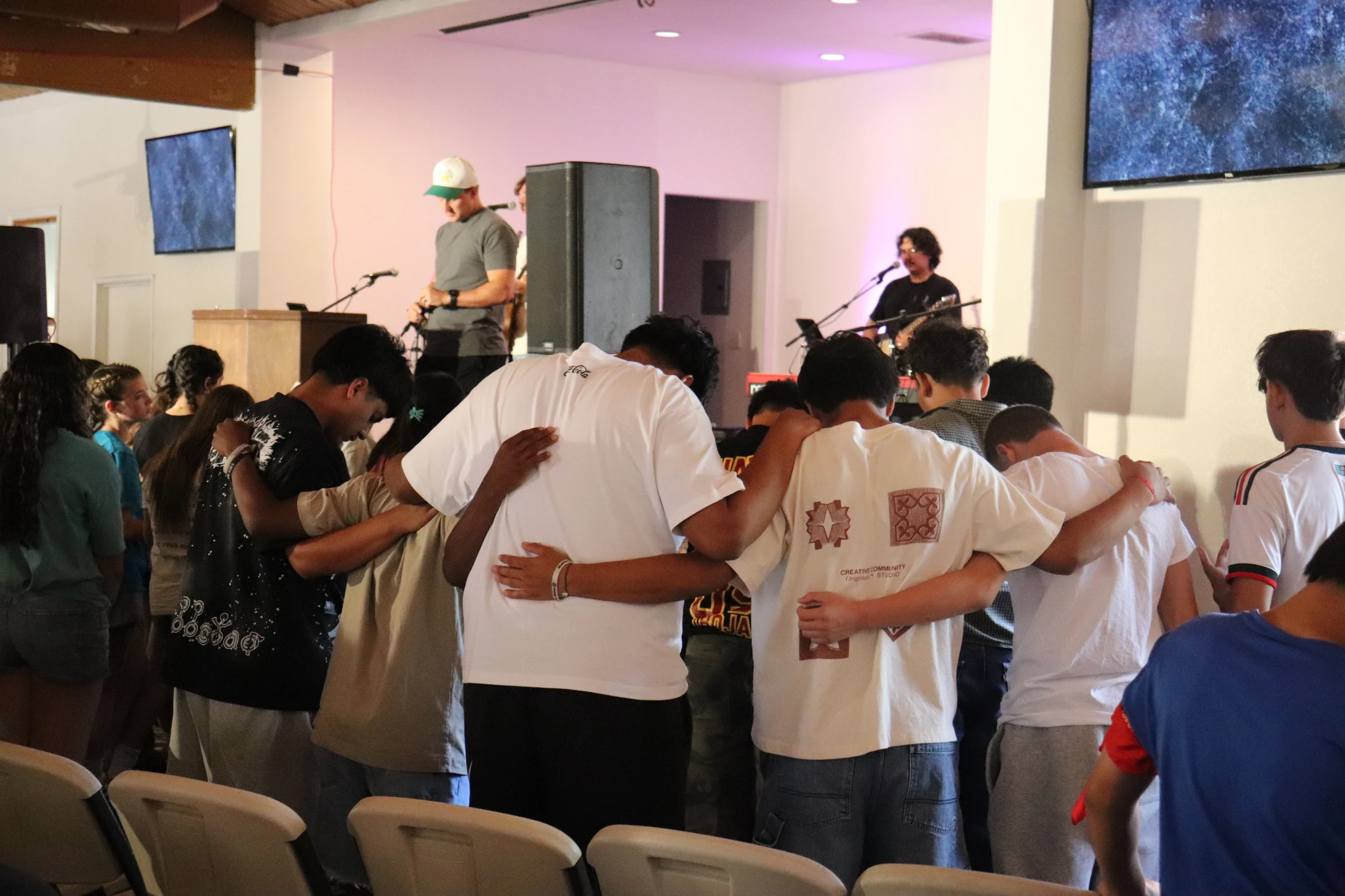 A group of young people stand with their arms around each other in prayer, facing a stage where a band is playing during a worship service.