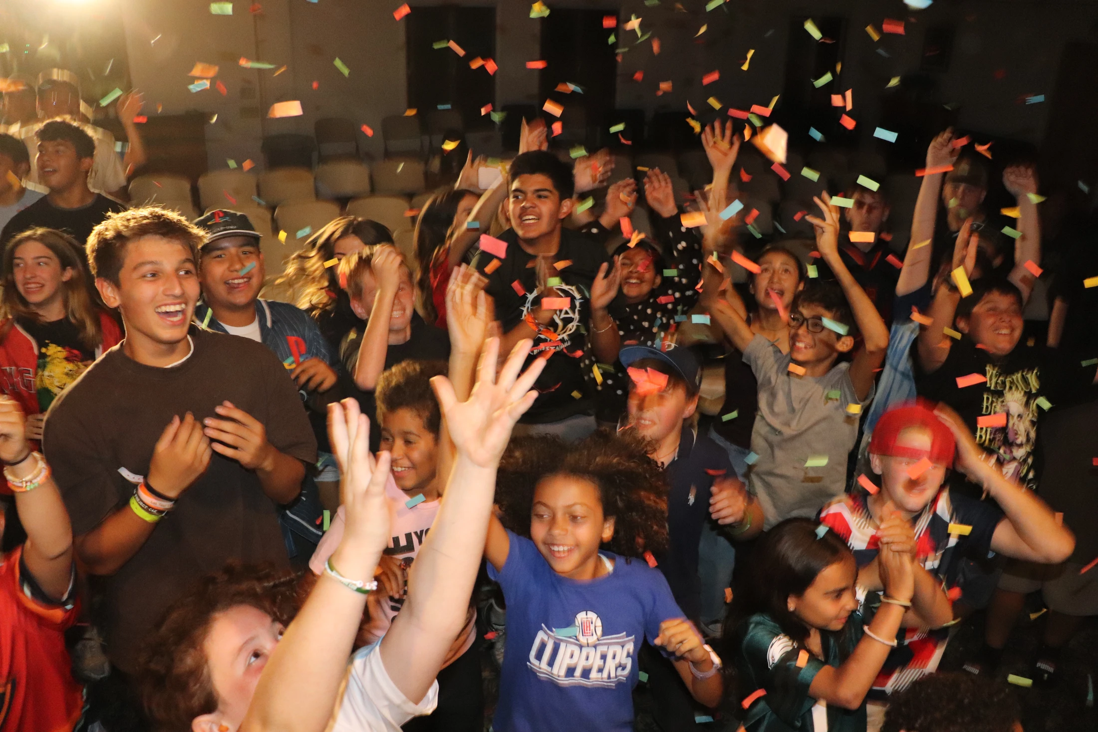 A large group of diverse young people cheer with their hands in the air as colorful confetti falls around them during a lively indoor celebration.