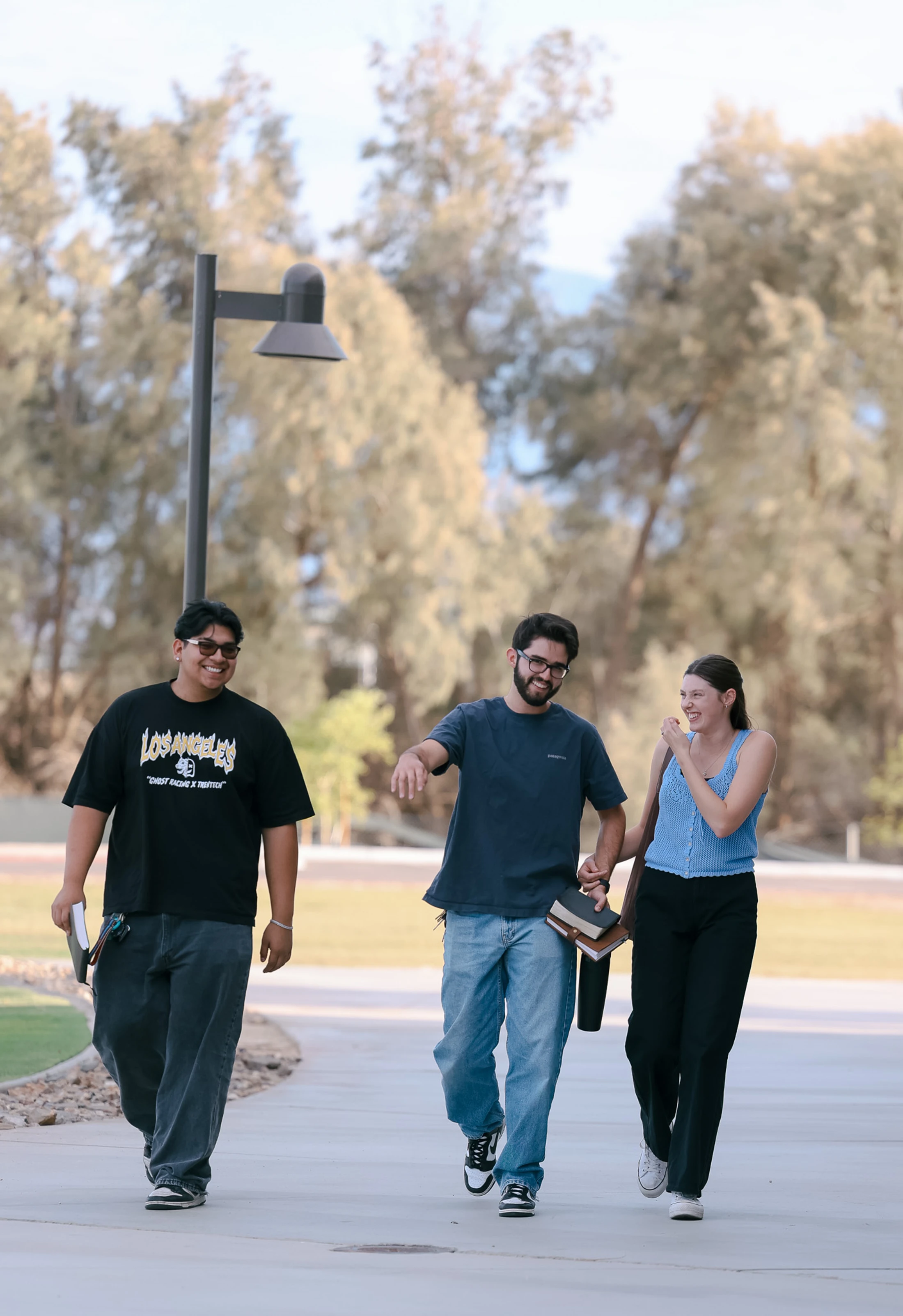 Three young students, two men and a woman, walk together on a campus path, smiling and chatting while carrying books on a sunny day.