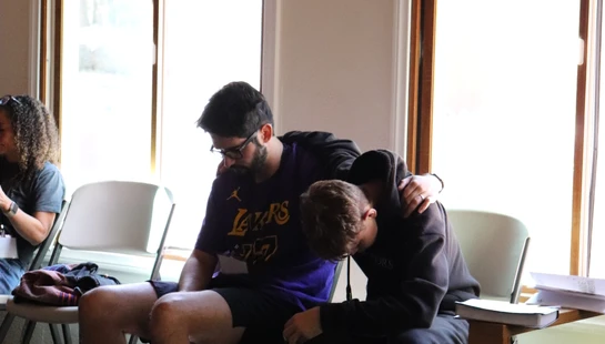 Two young men sit in chairs. One man in a Lakers shirt has his arm around another man who has his head bowed, offering comfort and support in a sunlit room.