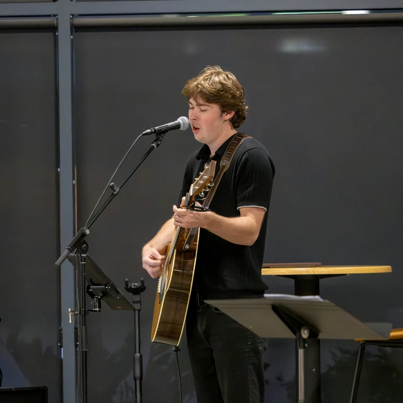 A young man with brown hair stands on a stage, playing an acoustic guitar and singing passionately into a microphone.