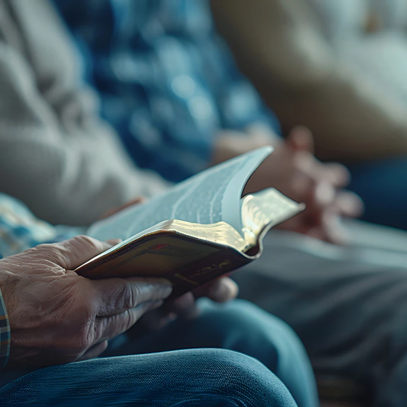 A close-up of an elderly man's hands holding an open Bible while sitting in a group. He is wearing a plaid shirt and the background is blurred.
