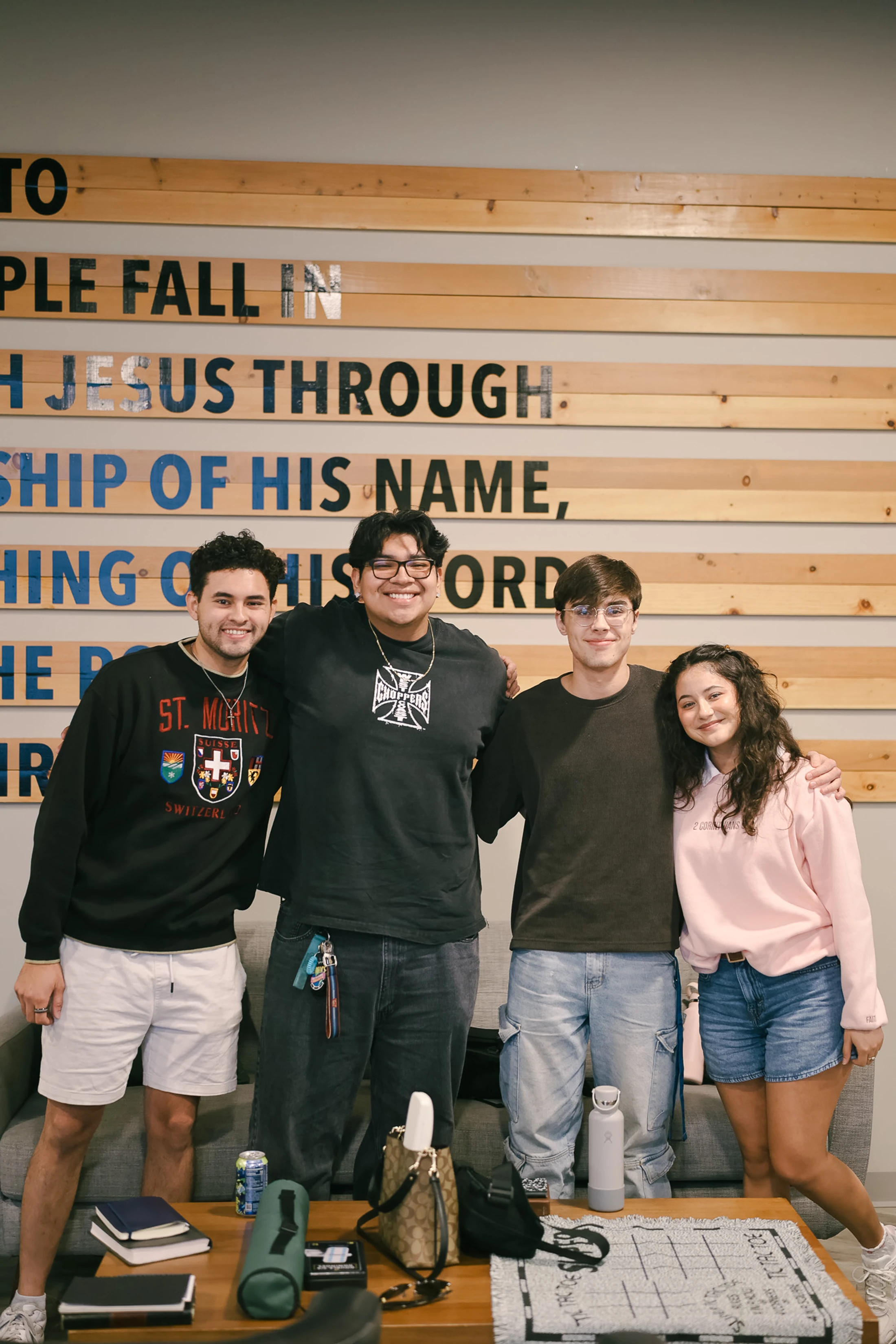 Four young adults, three men and one woman, stand together with their arms around each other, smiling at the camera in front of a wood-paneled wall.