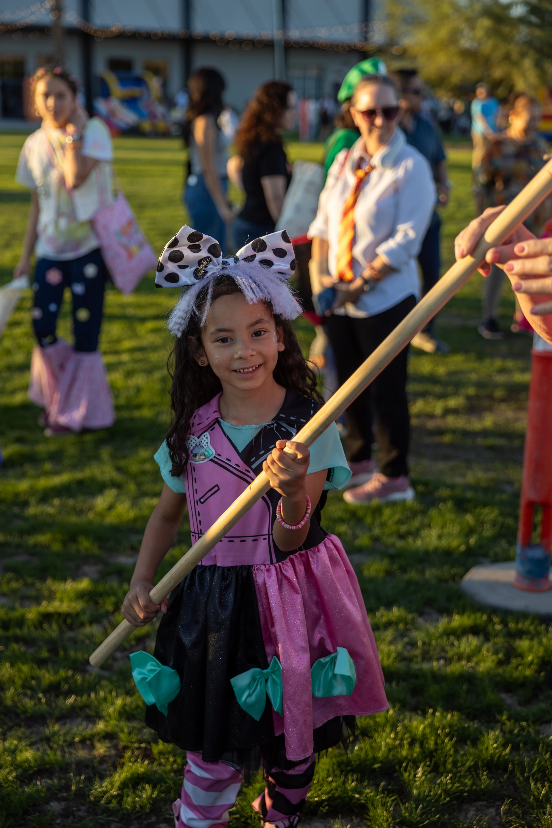 A young girl with a large polka-dot bow smiles at the camera while wearing a pink and black costume and holding a long wooden stick at a sunny outdoor event.