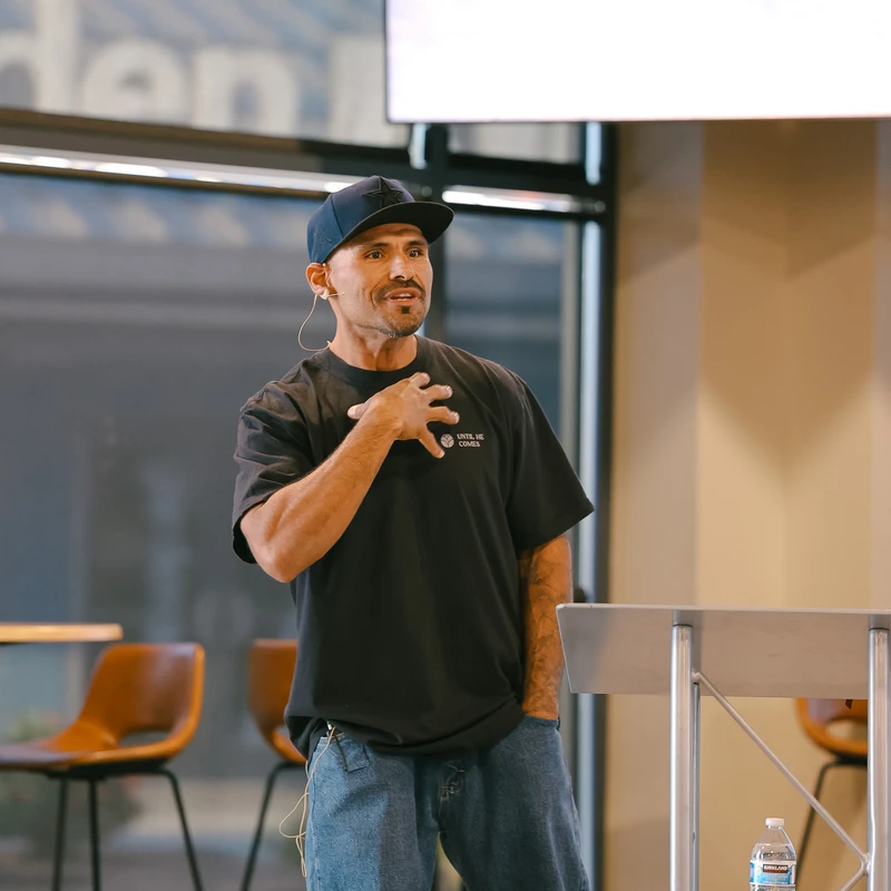 A man wearing a black cap and t-shirt stands behind a lectern, speaking with his hand on his chest. A screen behind him reads 'THE SELFLESS PHILIPPIANS'.