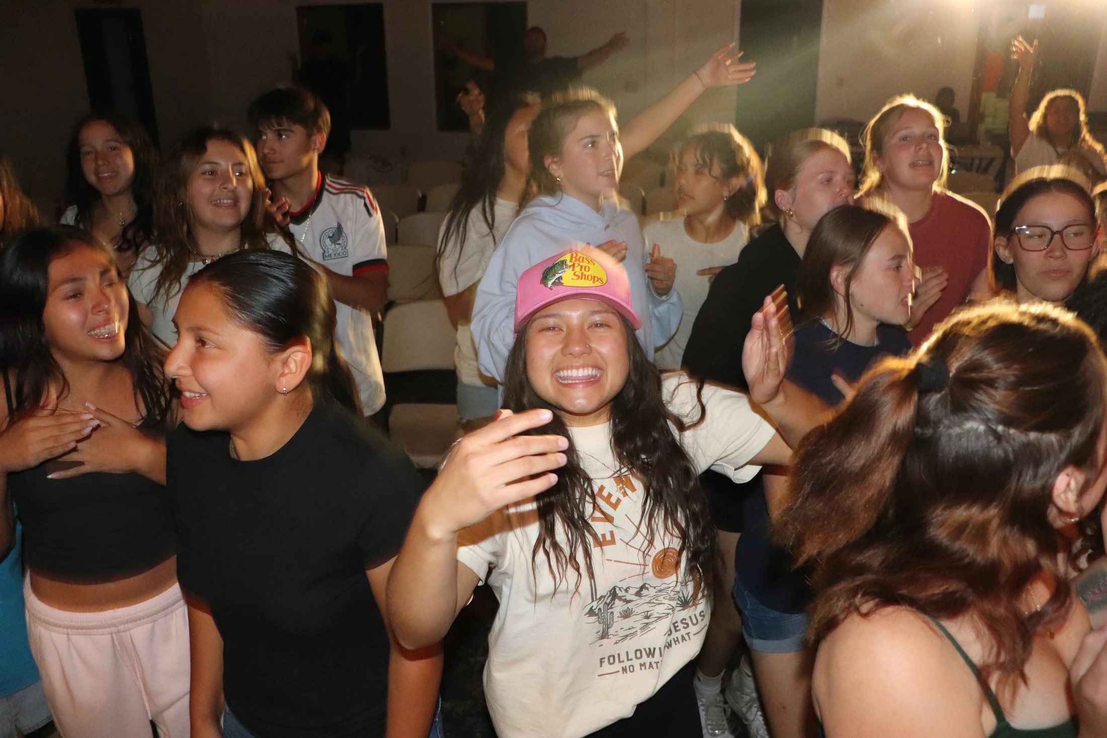 A large group of diverse teenagers and young adults sing and dance together in a warmly lit room. A woman in a pink hat smiles brightly at the camera.