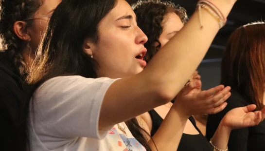 A young woman with dark hair wearing a Disney t-shirt sings with her eyes closed and hands raised in a group of people at an indoor gathering.