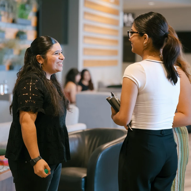 Two women stand facing each other, smiling and talking in a modern office lounge. One wears a black lace top, the other a white shirt and glasses.