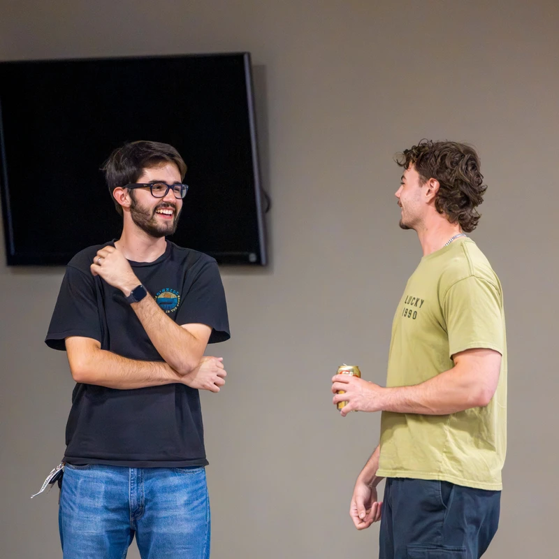 Two young men stand indoors talking. The man on the left, with glasses and a beard, smiles with his arms crossed. The other holds a can as they chat.