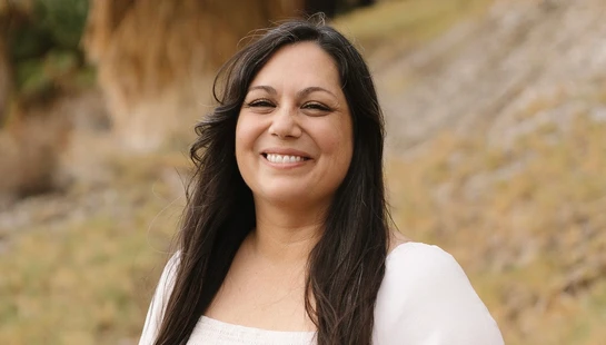 A portrait of a woman with long, dark hair smiling warmly at the camera. She is outdoors in a natural setting with trees and a hill in the background.