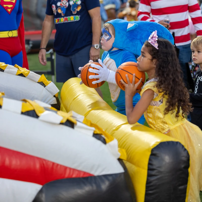Children in Sonic and princess costumes hold basketballs, preparing to play an inflatable game as adults in Superman and Waldo costumes look on.