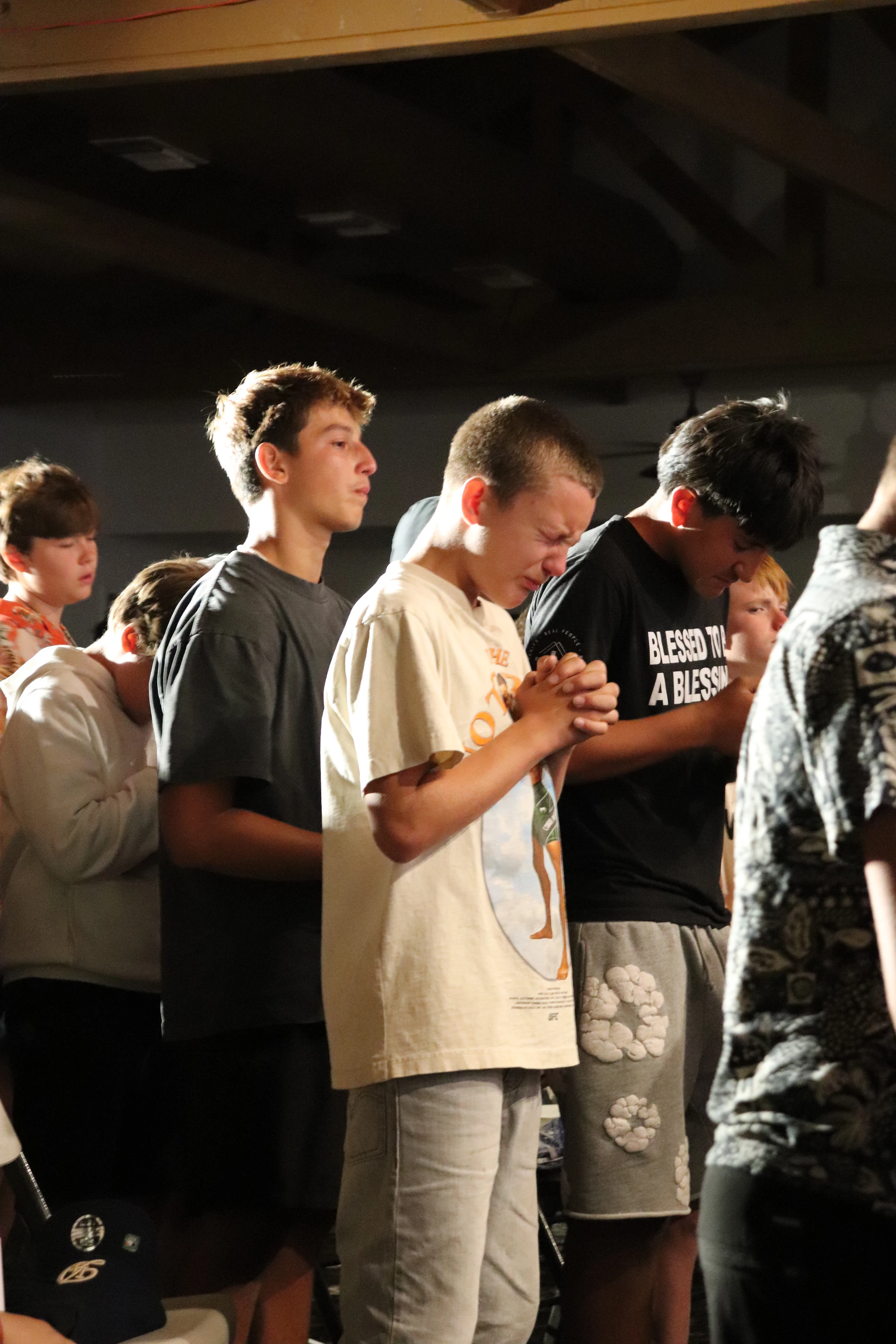 A group of teenage boys stand with their heads bowed in prayer. The boy in the center is visibly emotional, crying with his hands clasped together.