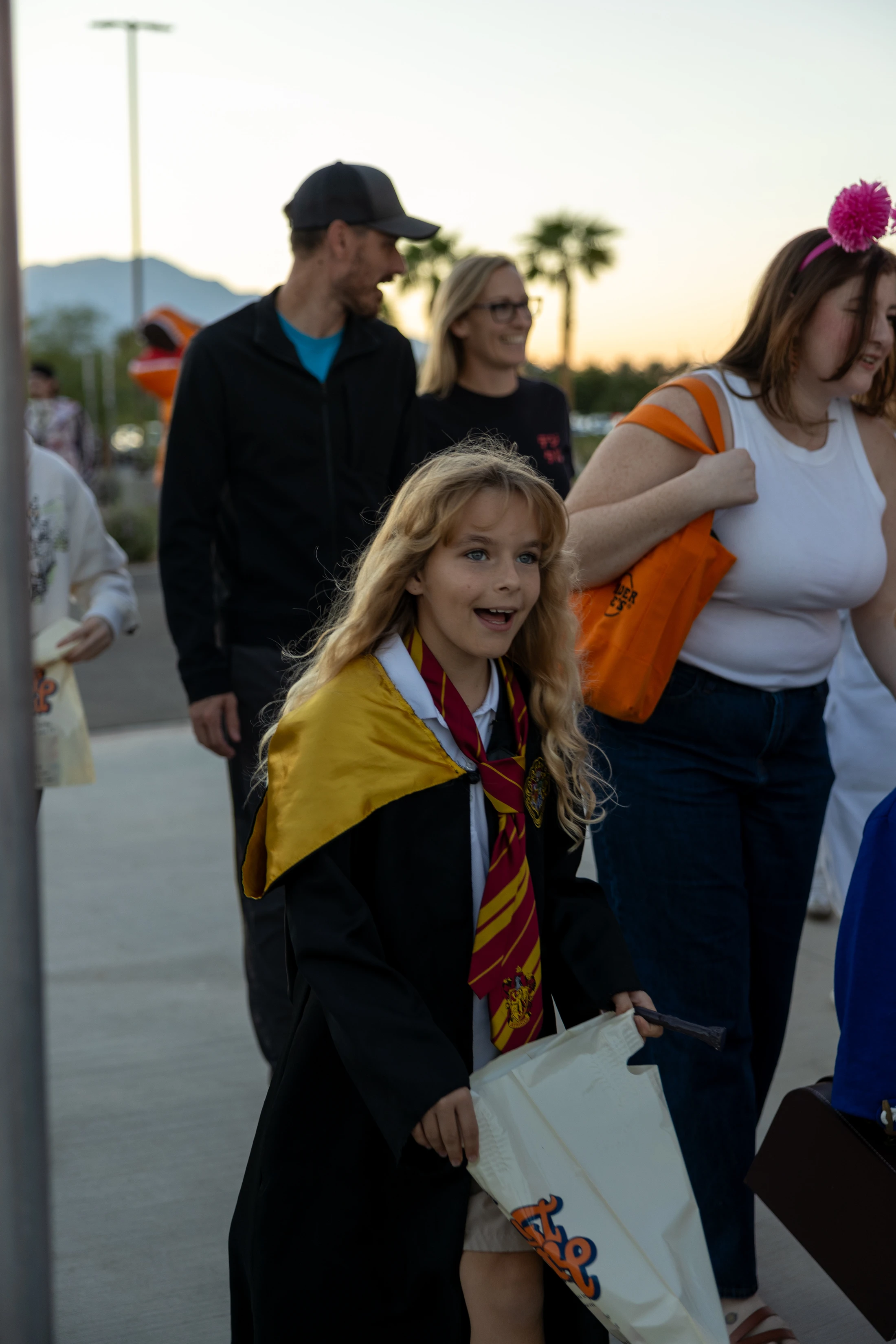 A happy young girl with long blonde hair dressed in a Harry Potter Gryffindor robe and tie walks outdoors with a look of pure excitement on her face.