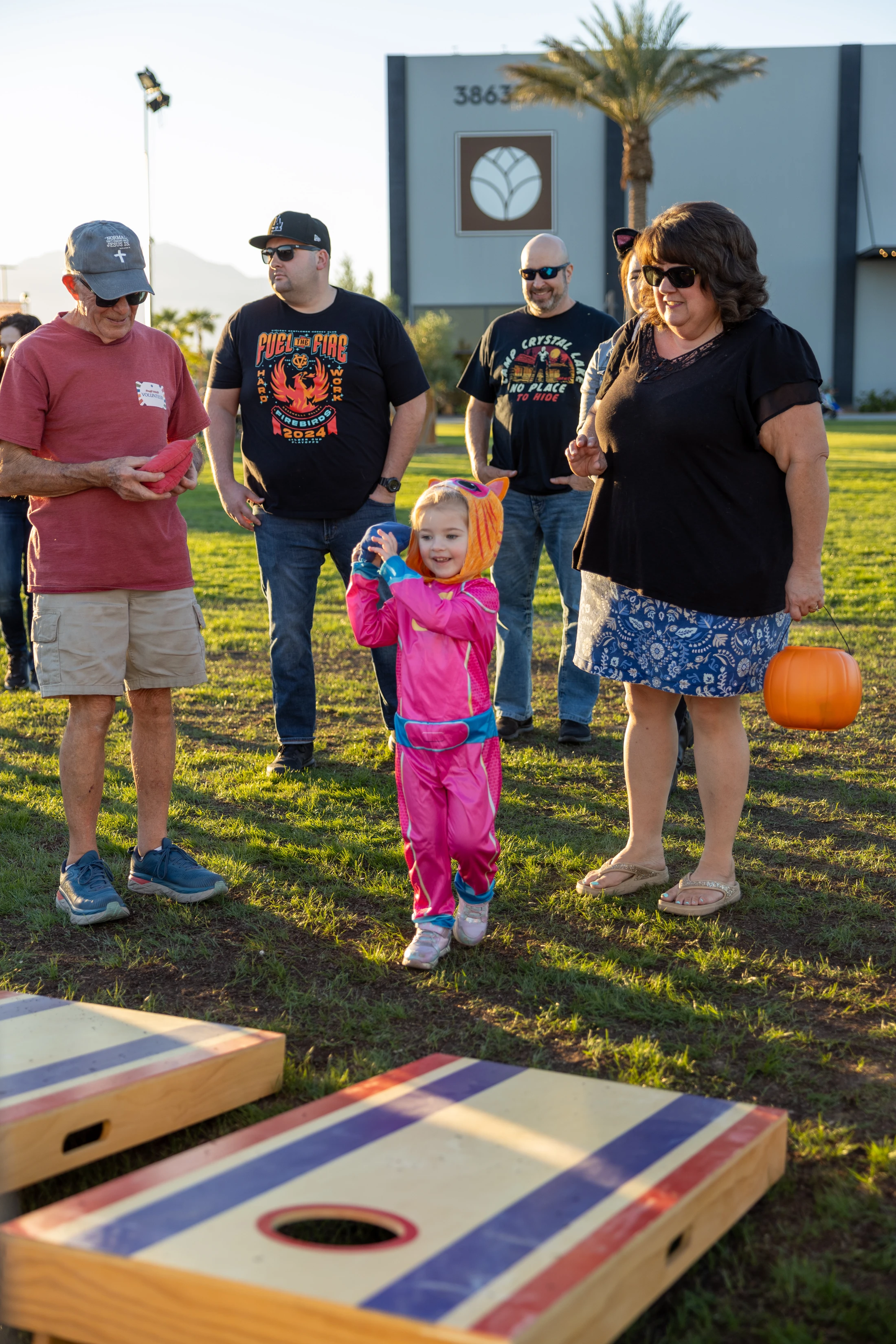 A young girl in a pink and orange costume smiles as she gets ready to toss a bean bag while playing cornhole on a grassy lawn with adults watching.