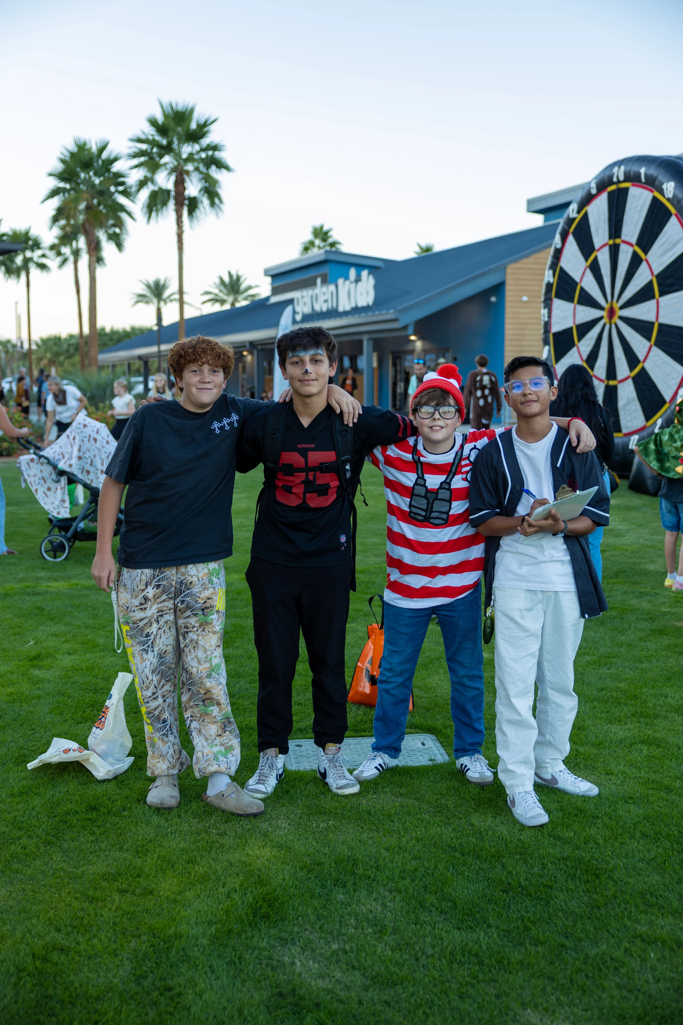 A group of four young boys in costumes, including one as Where's Waldo, stand with their arms around each other on a grassy lawn at an outdoor festival.