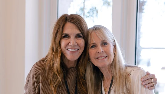 Two smiling women pose for a photo indoors in front of a window. The woman on the left has brown hair and the woman on the right has blonde hair.