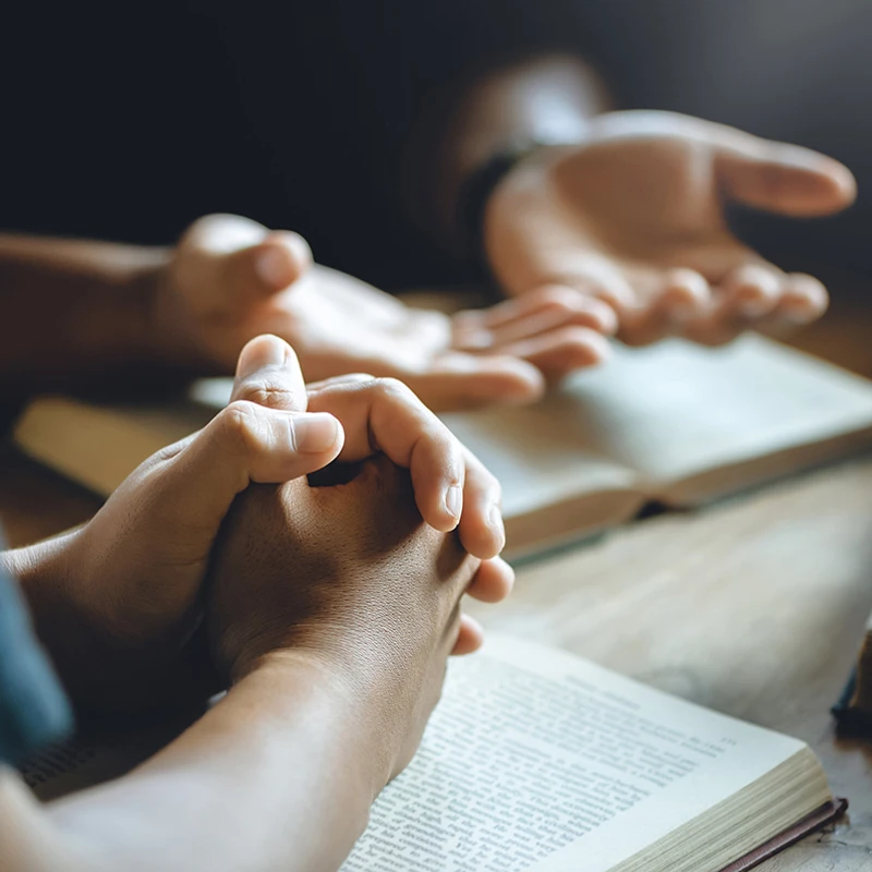 A close-up of people's hands resting on open Bibles at a wooden table. One person has their hands clasped while another gestures with open palms.