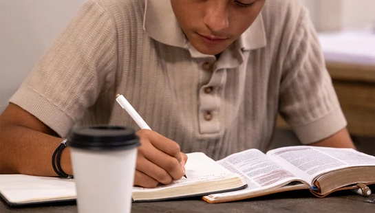 A young man in a beige polo shirt sits at a table, writing in a notebook next to an open book and a coffee cup, concentrating on his studies.