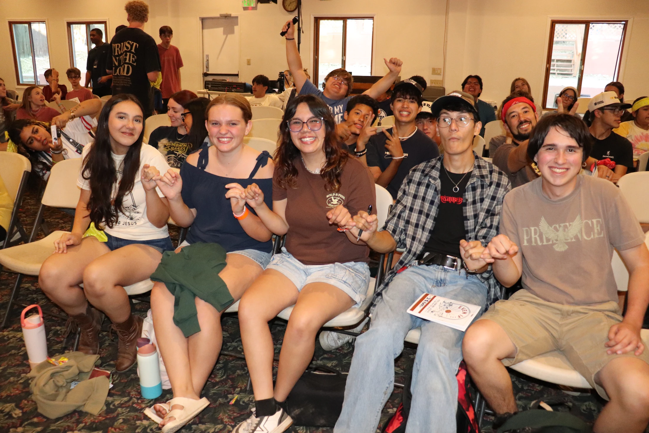 A large group of diverse teenagers sits in rows of chairs, smiling for a photo. The five young people in the front row pose together, linking their hands.