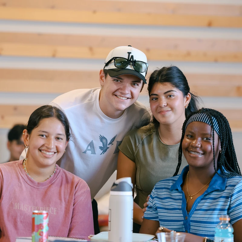 A diverse group of four students smile for the camera. A young man stands behind three young women sitting at a classroom table with books and drinks.