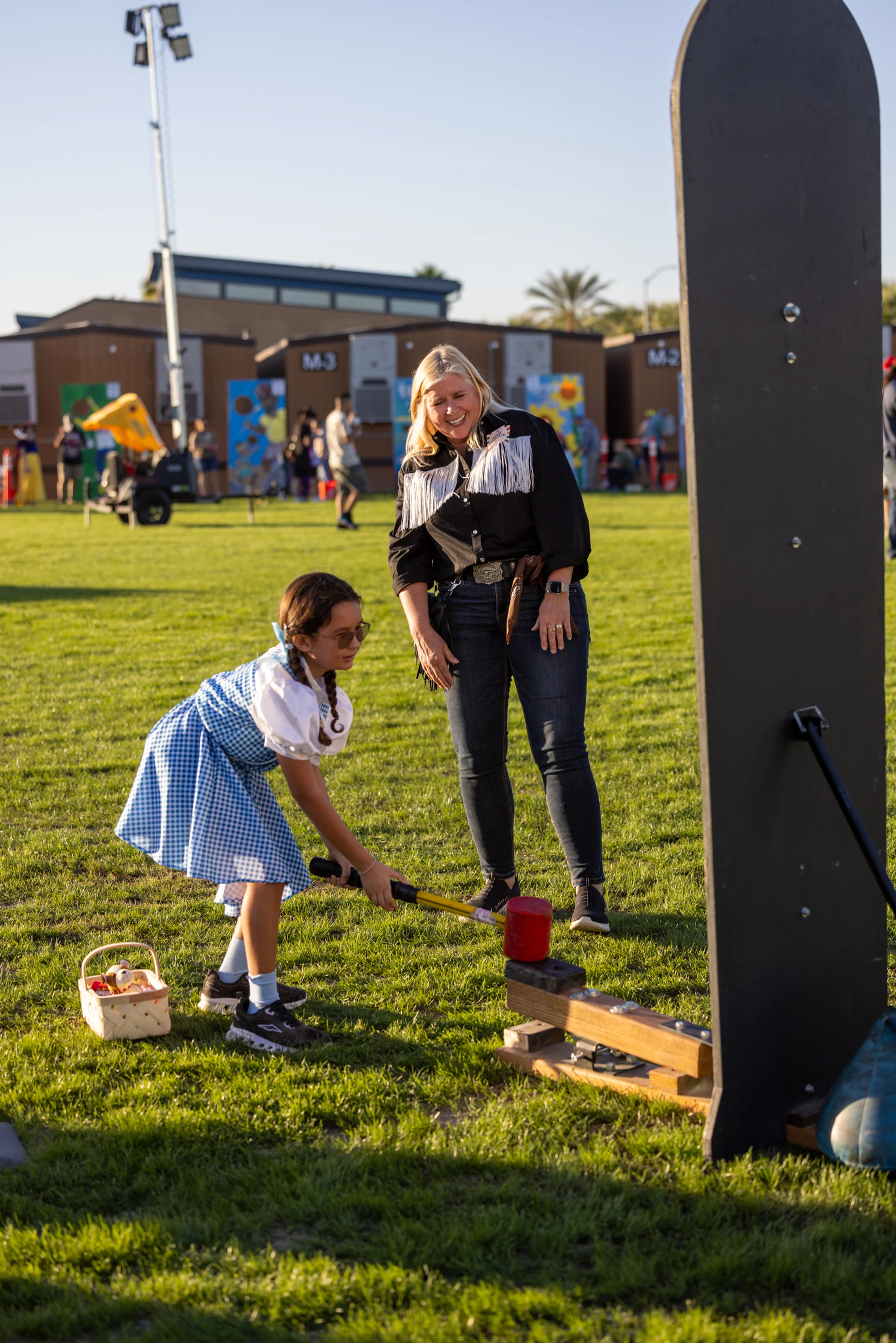 A young girl dressed as Dorothy from The Wizard of Oz swings a large mallet at a high-striker carnival game as a woman in a western shirt watches and smiles.