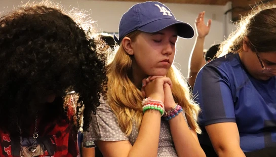 A group of young people stand with heads bowed. A girl in the center wears a baseball cap with her eyes closed and hands clasped together in prayer.