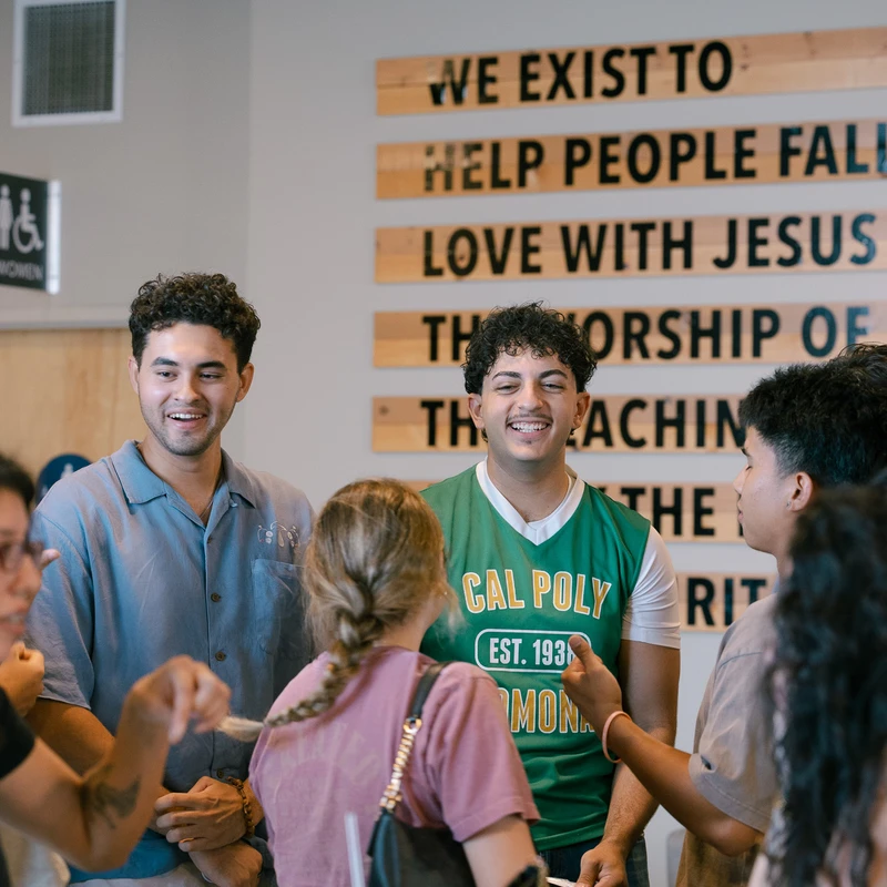 A group of diverse young adults smiling and talking together indoors in front of a wall sign that reads, 'WE EXIST TO HELP PEOPLE FALL IN LOVE WITH JESUS'.
