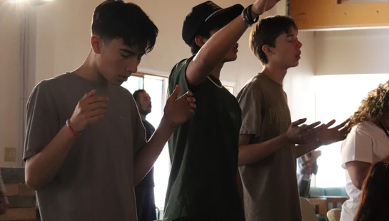 Three young men stand indoors in front of chairs, with their eyes closed and hands raised in a gesture of worship or prayer.